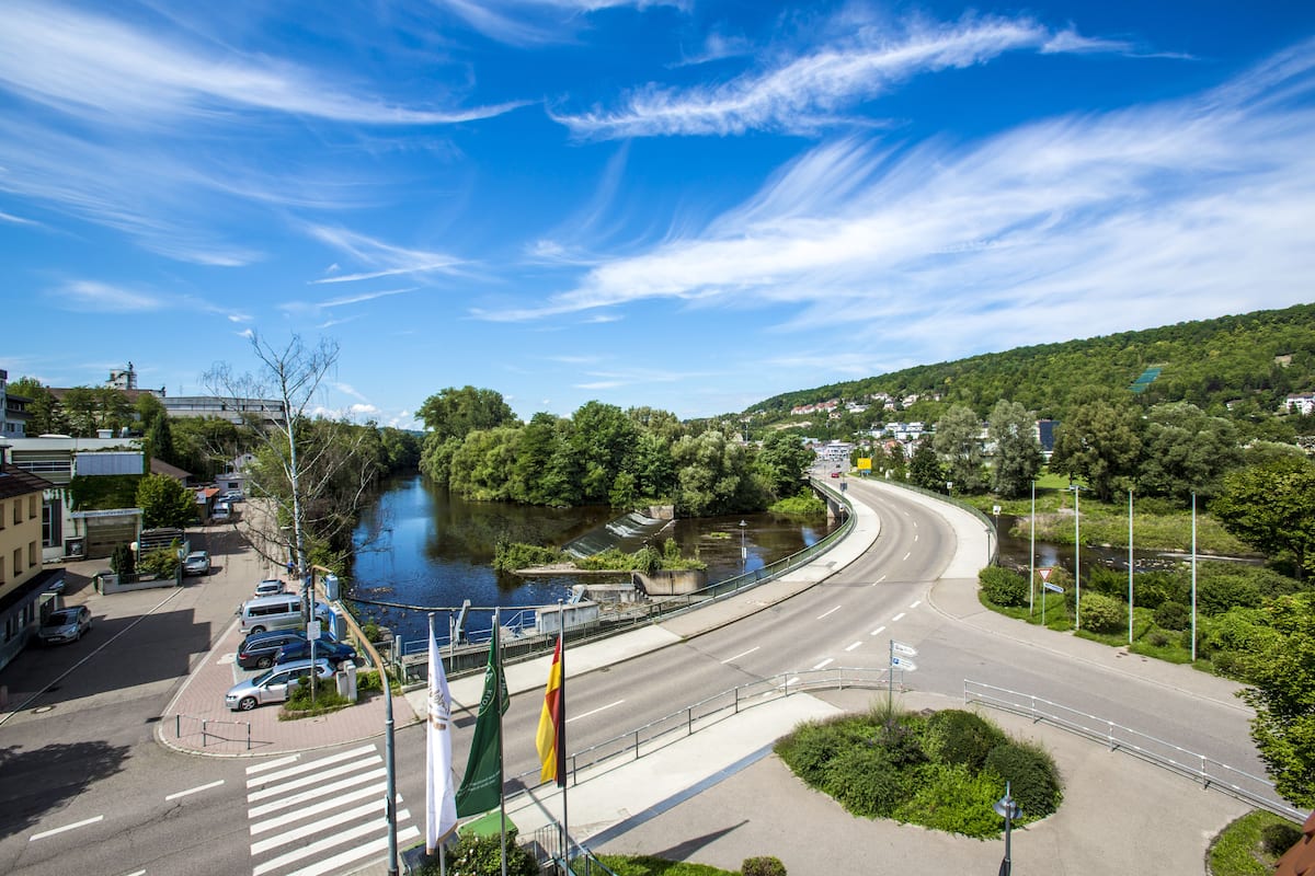 A river running alongside a road with a bridge, greenery, and buildings in the background.