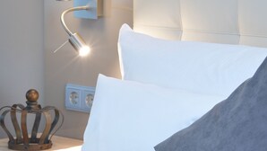 Una cama bien hecha con almohadas blancas y grises, una lámpara de noche y una corona decorativa en la mesita de noche.