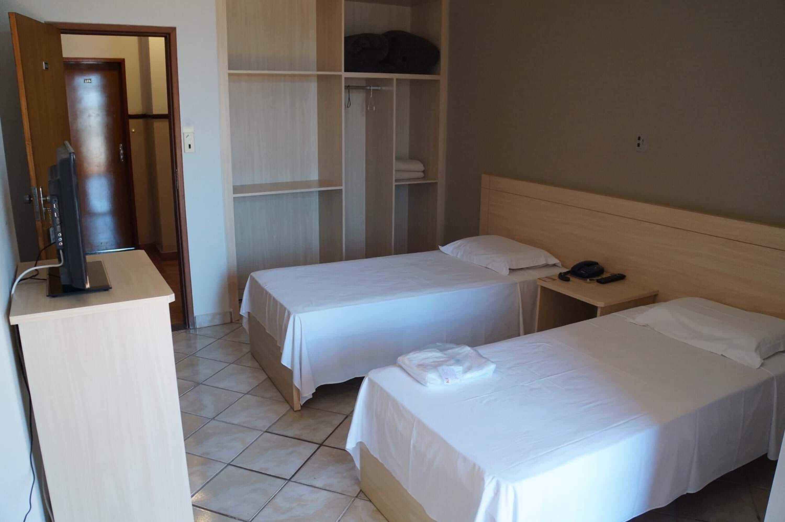 Ett hotellrum med två separata sängar, ett skrivbord med en telefon, en garderob och en tv som står på ett stativ.