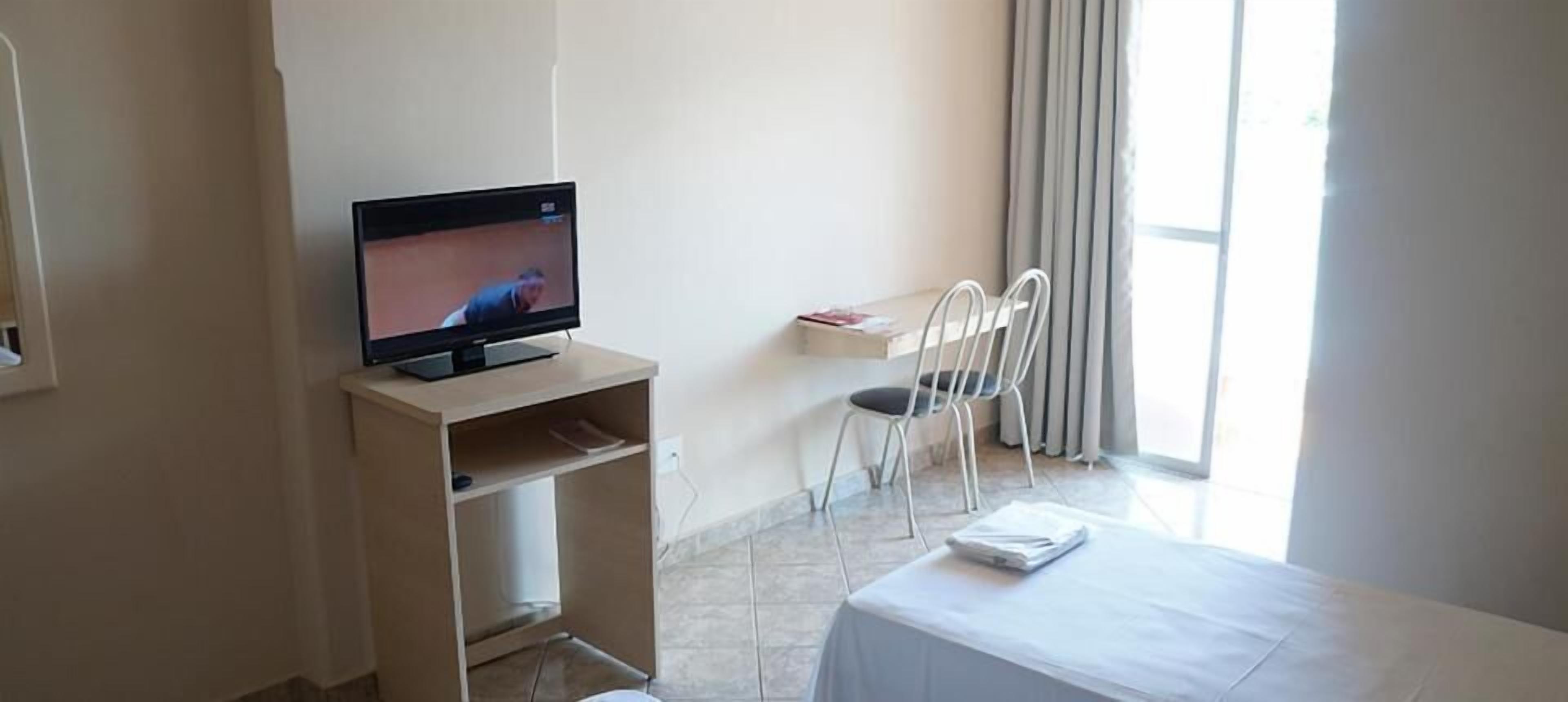 Habitación de hotel con una televisión sobre un mueble bajo, una mesita con mantel blanco, una silla y una ventana con cortinas.
