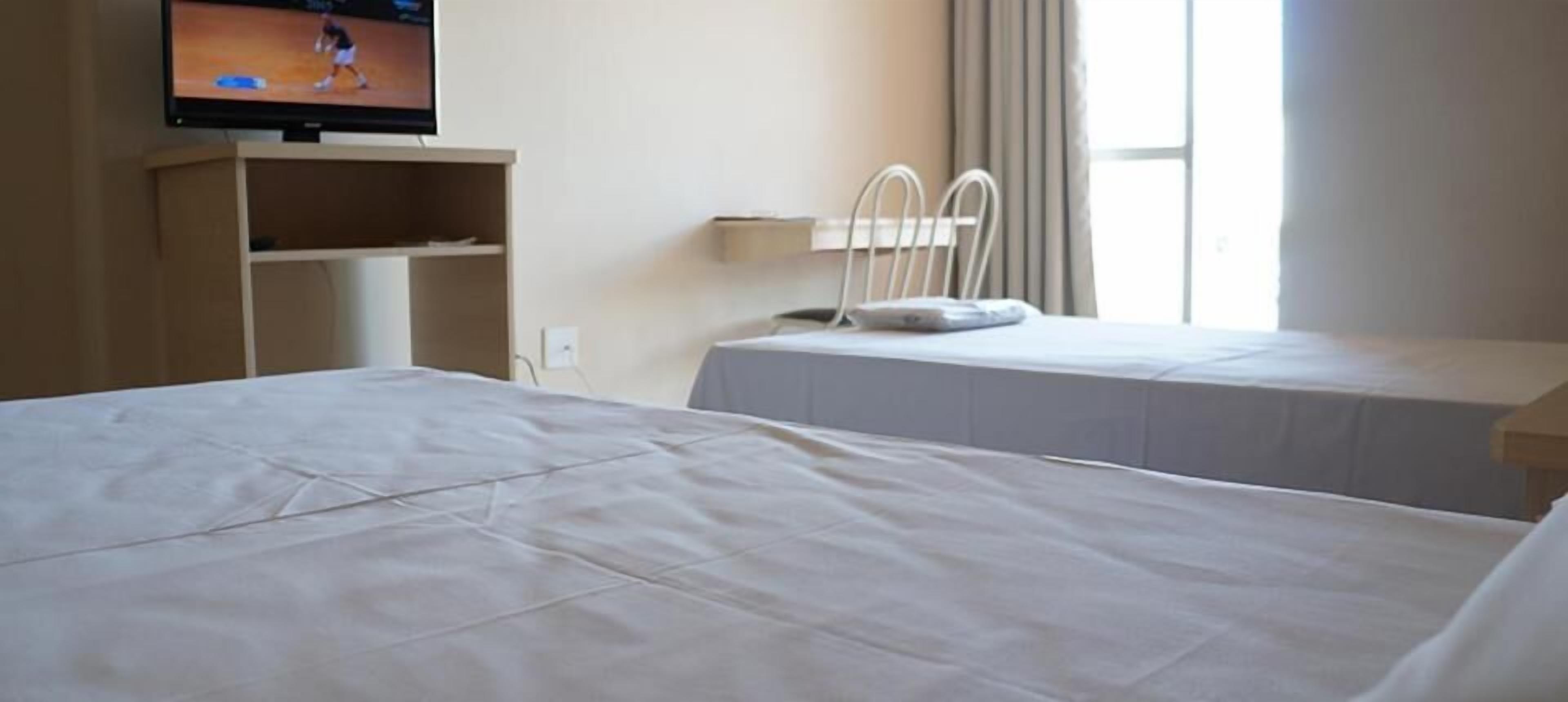 Ein Hotelzimmer mit einem großen Bett, einem an der Wand befestigten Fernseher, einem Nachttisch und einem Fenster mit Vorhängen.