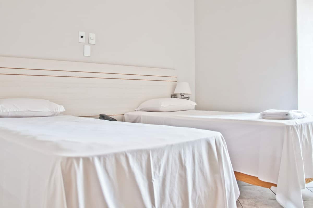 A hotel room with two single beds, a bedside table with a lamp, and a wall-mounted light switch.
