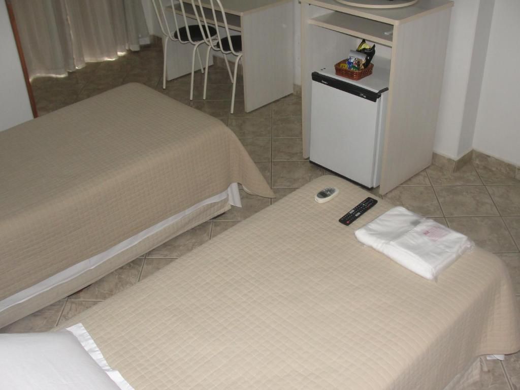 A hotel room with two beds, a desk, a chair, a refrigerator, and a remote control on the bed.