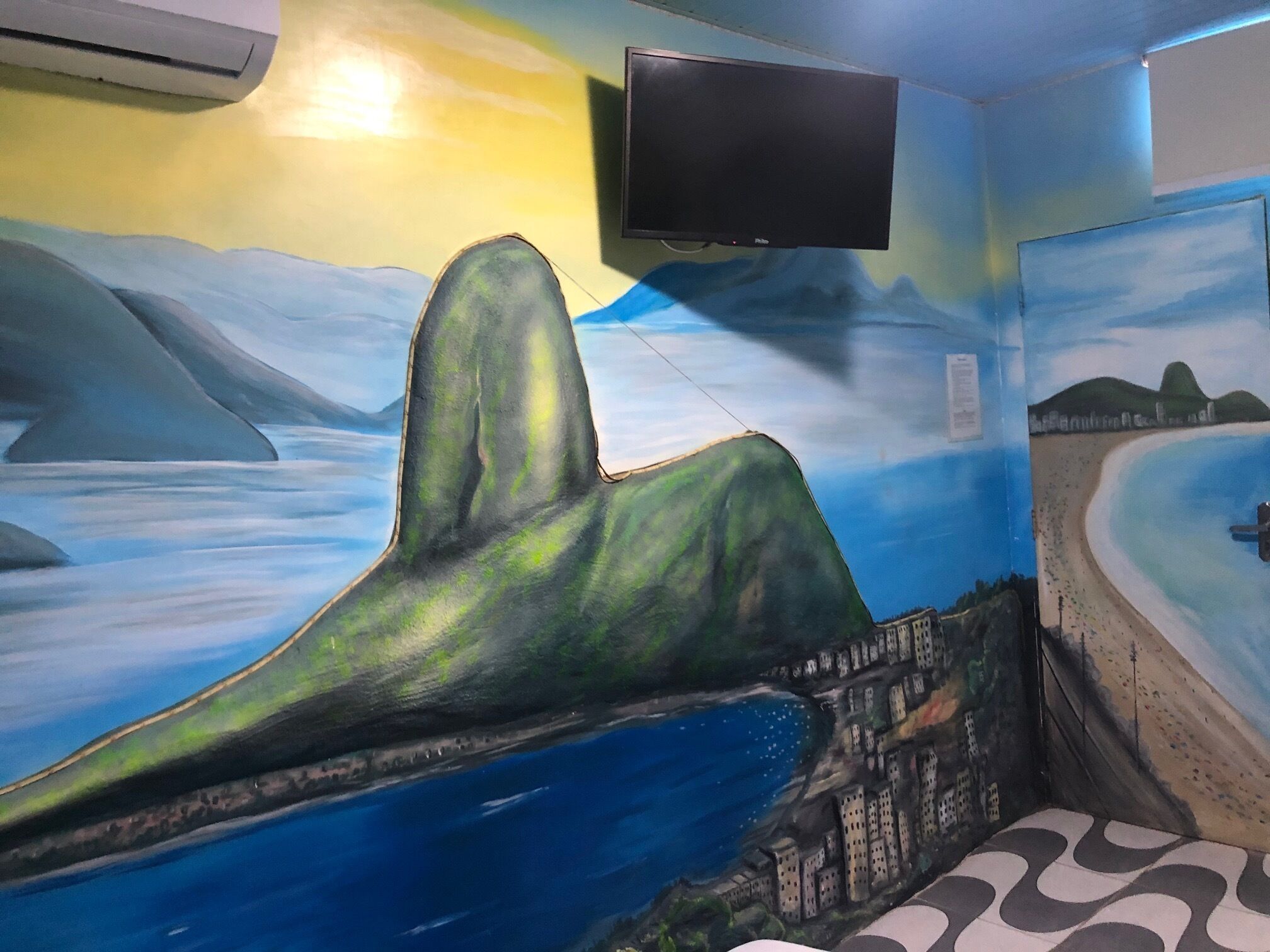 Quarto com um mural representando uma paisagem costeira, uma TV de tela plana na parede e um tapete estampado no chão.