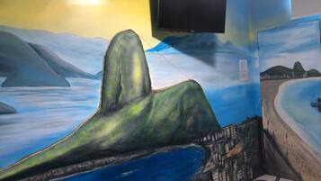 Quarto com um mural representando uma paisagem costeira, uma TV de tela plana na parede e um tapete estampado no chão.