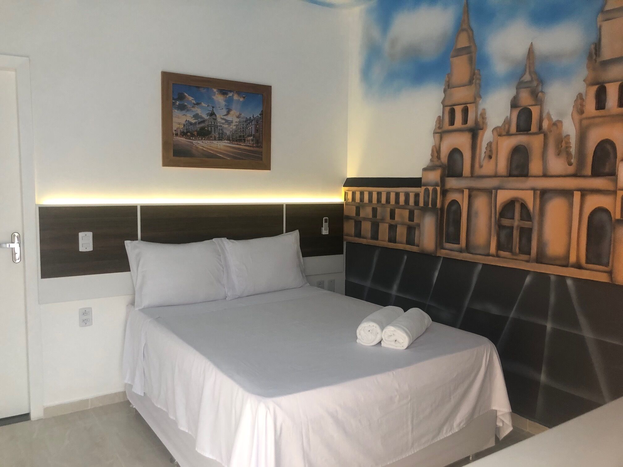 A hotel room with a bed, a headboard, a painting, and a mural on the wall.