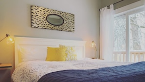 A bed with a white headboard, a yellow pillow, and a dark blue quilt. There is a wall-mounted mirror above the bed and a window with a view of trees. - Joanna Gaines would stay here! (Sandpoint)