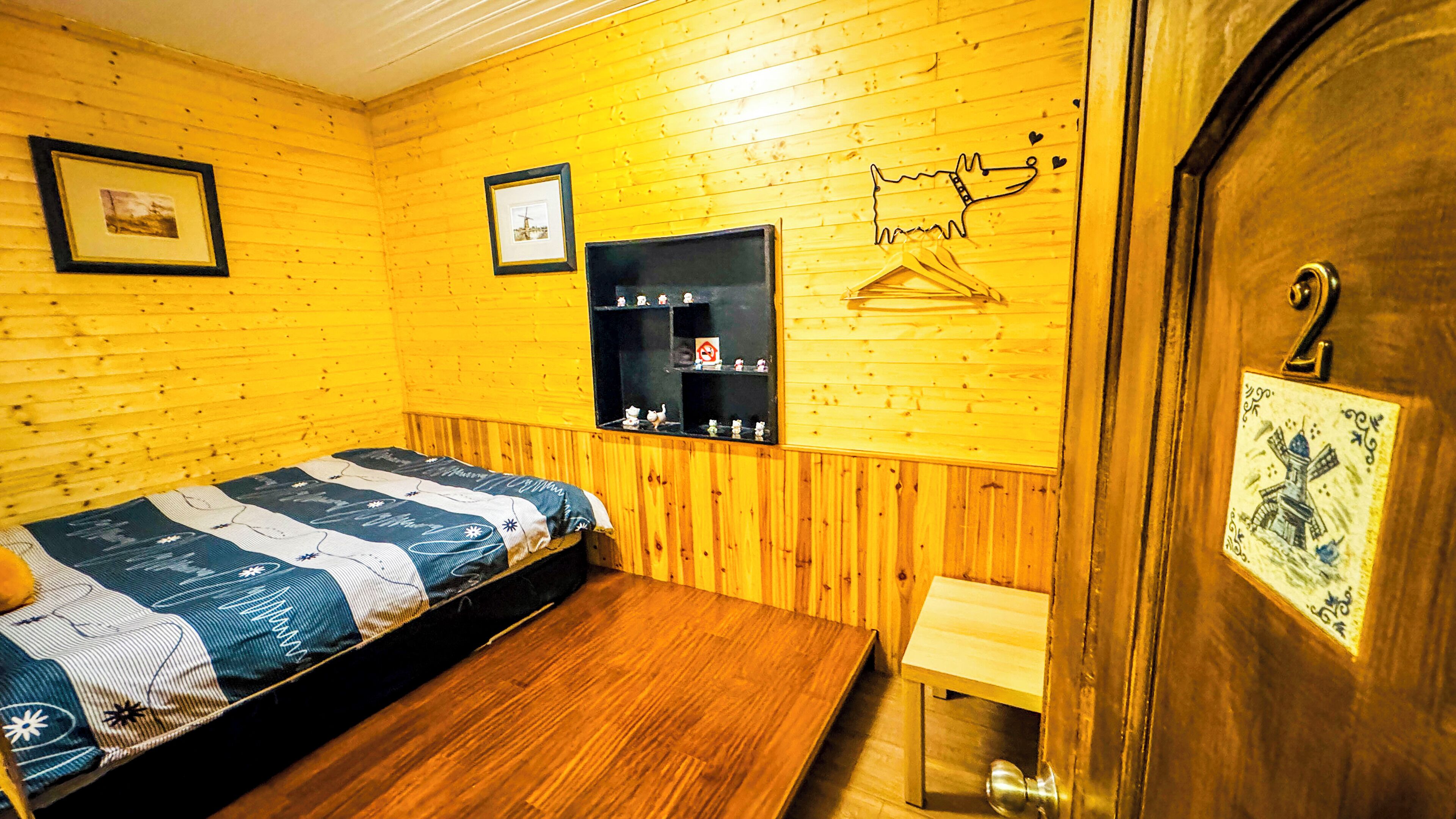 A room with wooden walls, a bed with a striped bedspread, a small bench, a shelf with figurines, and a door with a decorative plate.