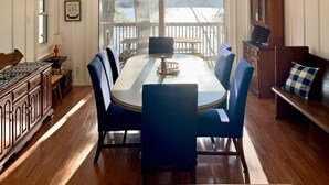 A dining room with a large wooden table, blue chairs, a ceiling fan, a wall clock, and a view through large windows.
