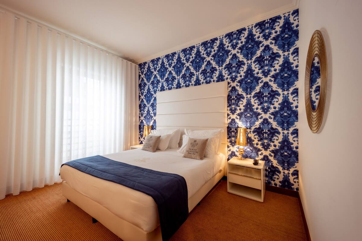 A hotel room with a large bed, white headboard, and a blue patterned wall.