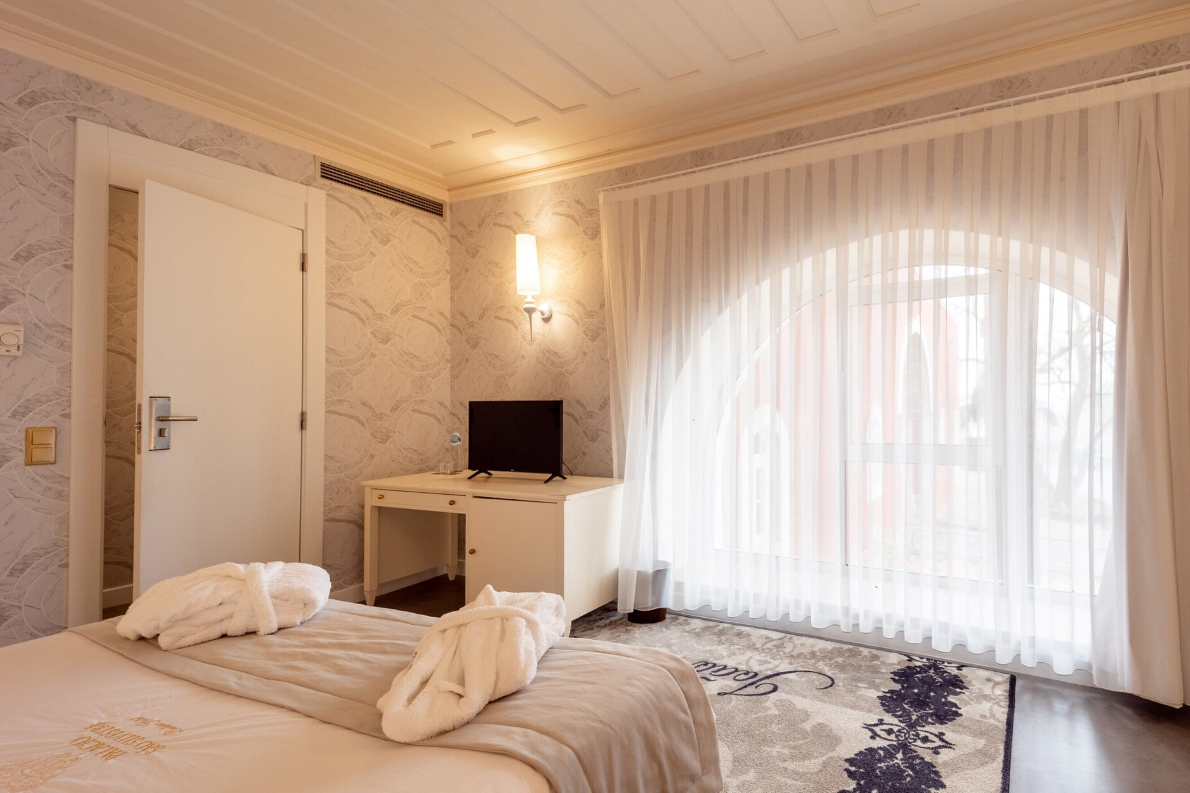 A hotel room with a bed, a desk, a TV, and a window with sheer curtains.
