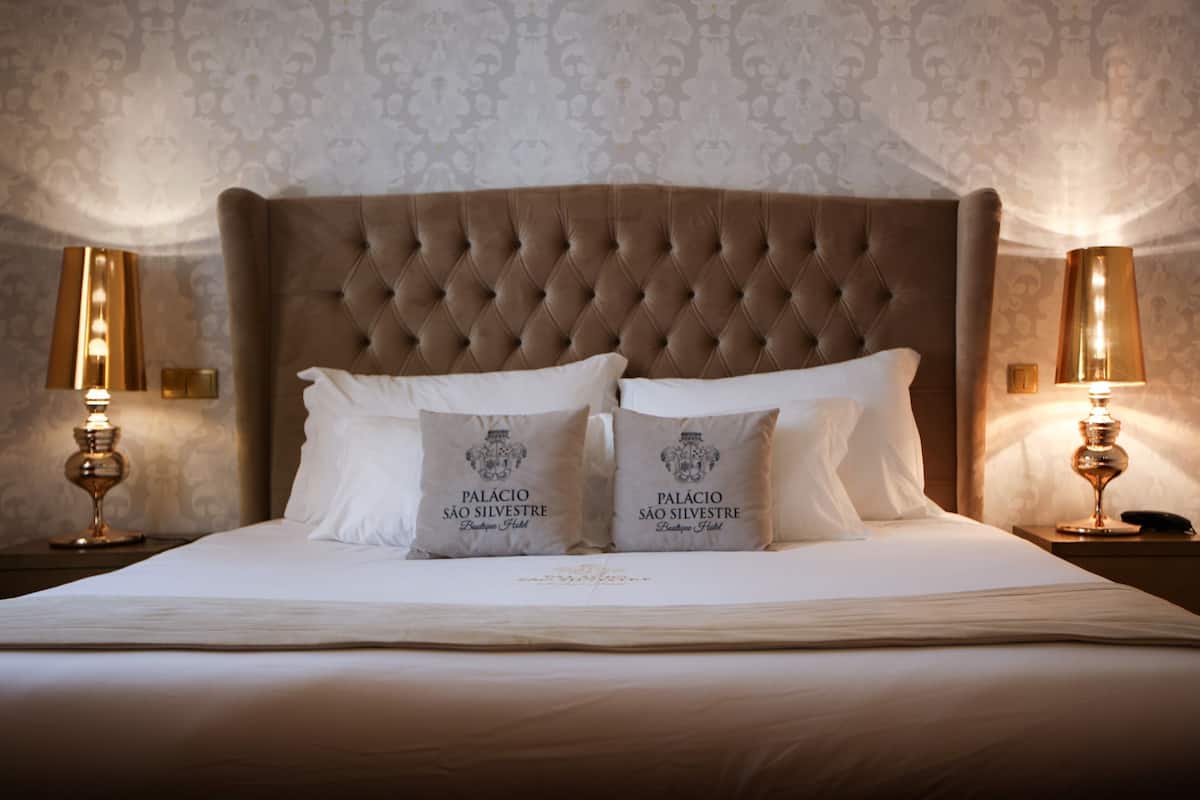 A neatly made bed with two pillows, each with a logo, in a room with patterned wallpaper and two bedside lamps.