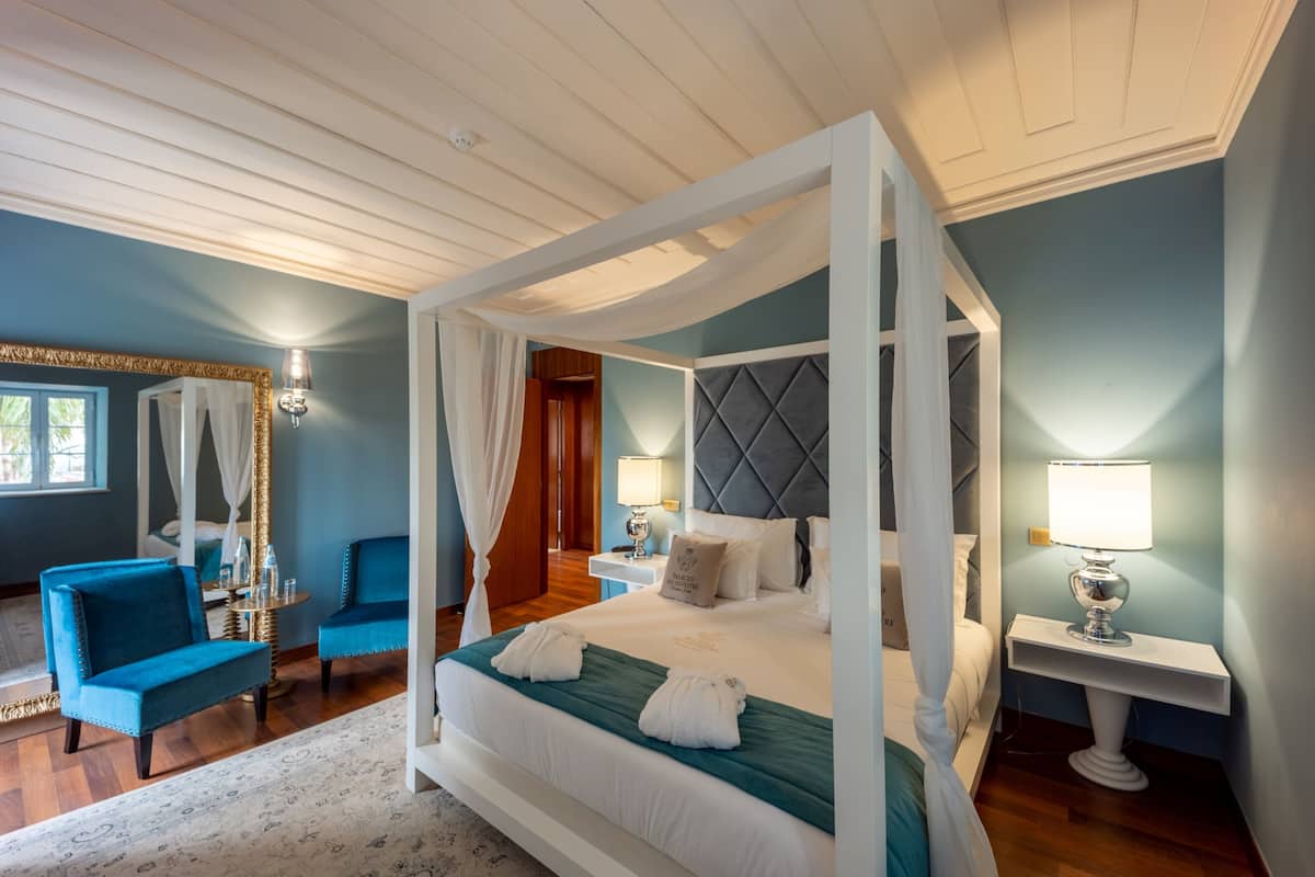 A four-poster bed with white linens, a teal accent bedspread, and two white towels on the bed. There are two bedside tables with lamps, a blue armchair, and a wooden nightstand with a mirror.