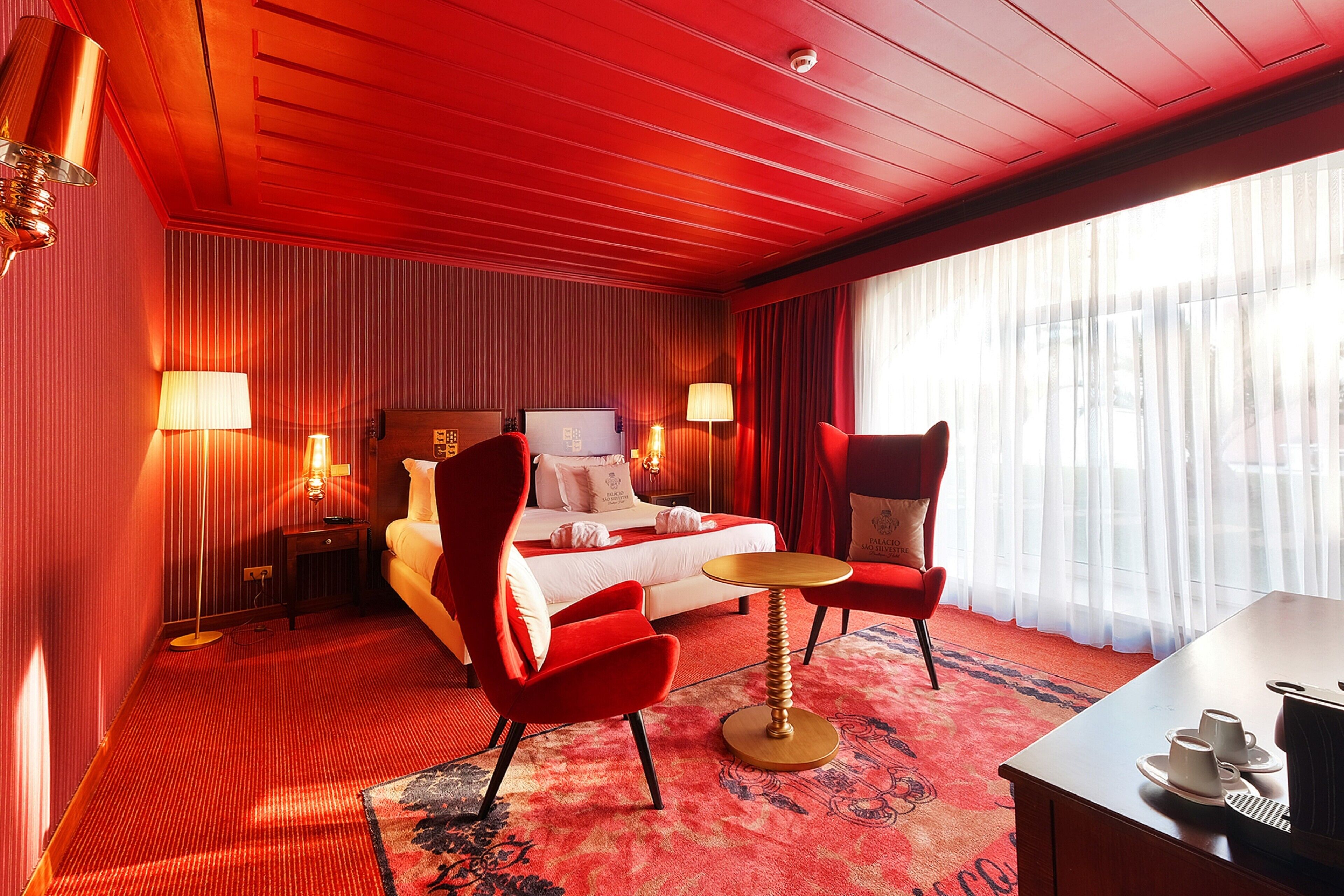 A hotel room with a red theme, featuring a bed, two red chairs, a small table, and a desk with cups and a kettle.