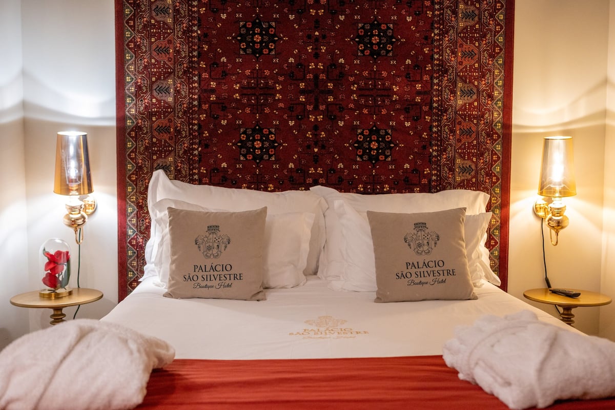 A hotel room with a bed, two pillows with the hotel's logo, a red bedspread, and two bedside lamps.