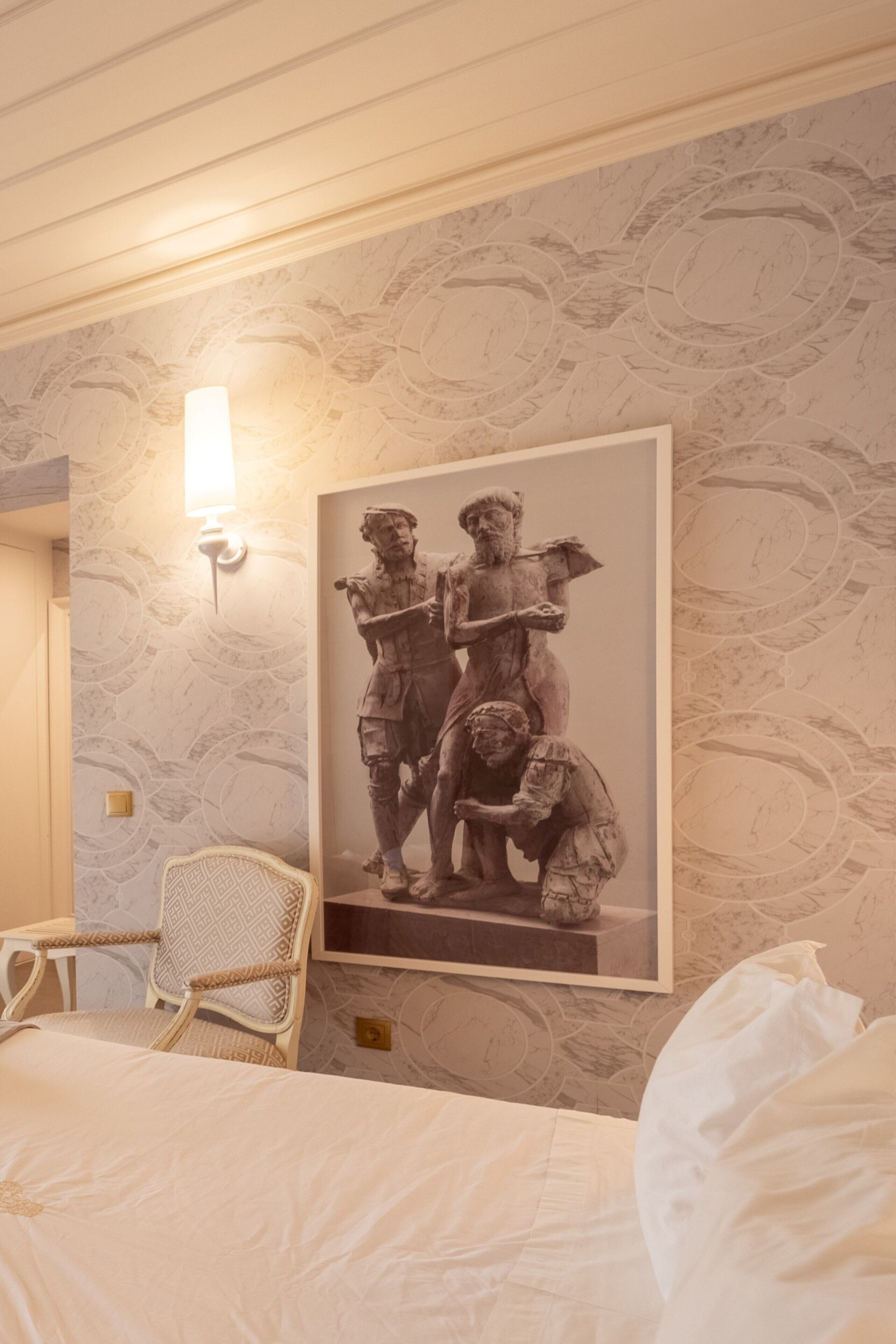 Une chambre d’hôtel comprenant un lit, une chaise, une lampe fixée au mur et une œuvre d’art encadrée représentant une scène historique.