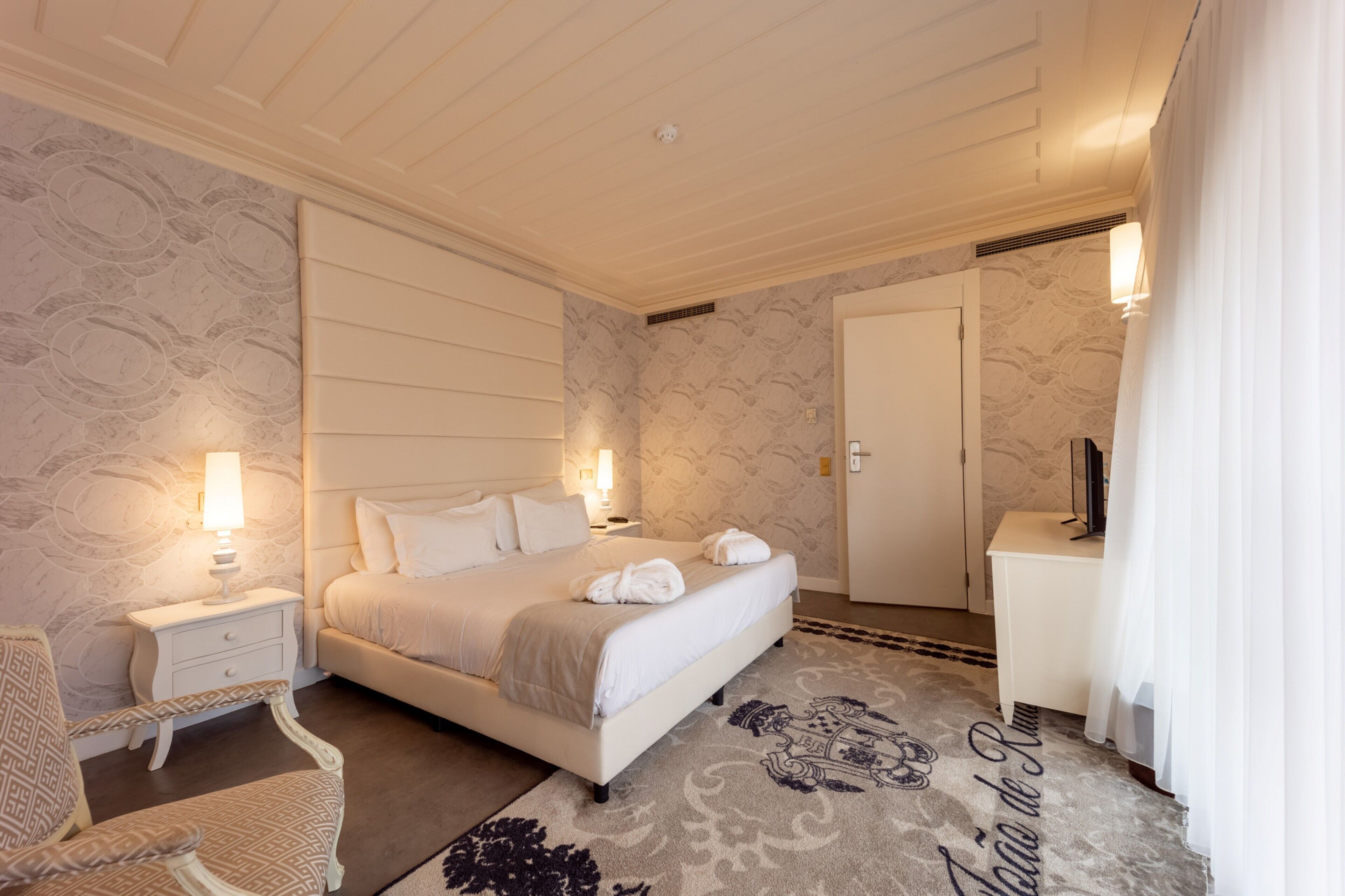 Quarto de hotel com uma cama grande, mesas de cabeceira com luminárias, uma cadeira, uma escrivaninha e um guarda-roupa.