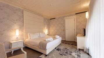Quarto de hotel com uma cama grande, mesas de cabeceira com luminárias, uma cadeira, uma escrivaninha e um guarda-roupa.