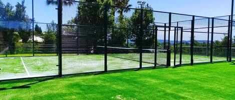 Un court de tennis entouré d’une clôture métallique, éclairé par des lampes solaires, et bordé d’arbres, avec un ciel dégagé au-dessus.