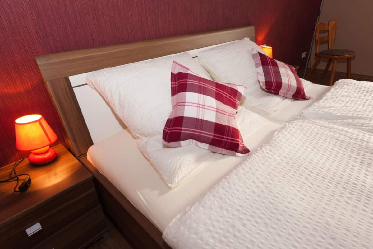 A neatly made bed with white bedding and two red and white checkered pillows. There is a bedside lamp on the left side of the bed, and a wooden chair is visible in the background.