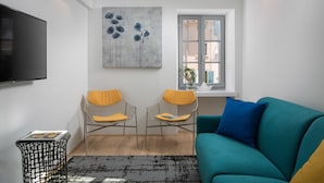A modern living room with a teal sofa, two wireframe chairs with yellow cushions, a black TV, a gray abstract painting, and a window with a view. - Alla Pescheria (Rovinj)