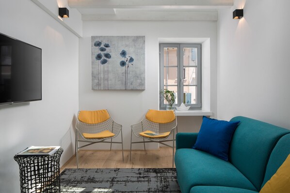 Uma sala de estar moderna com um sofá azul-turquesa, duas cadeiras de metal com almofadas amarelas, uma TV preta, um quadro abstrato cinzento e uma janela com vista.