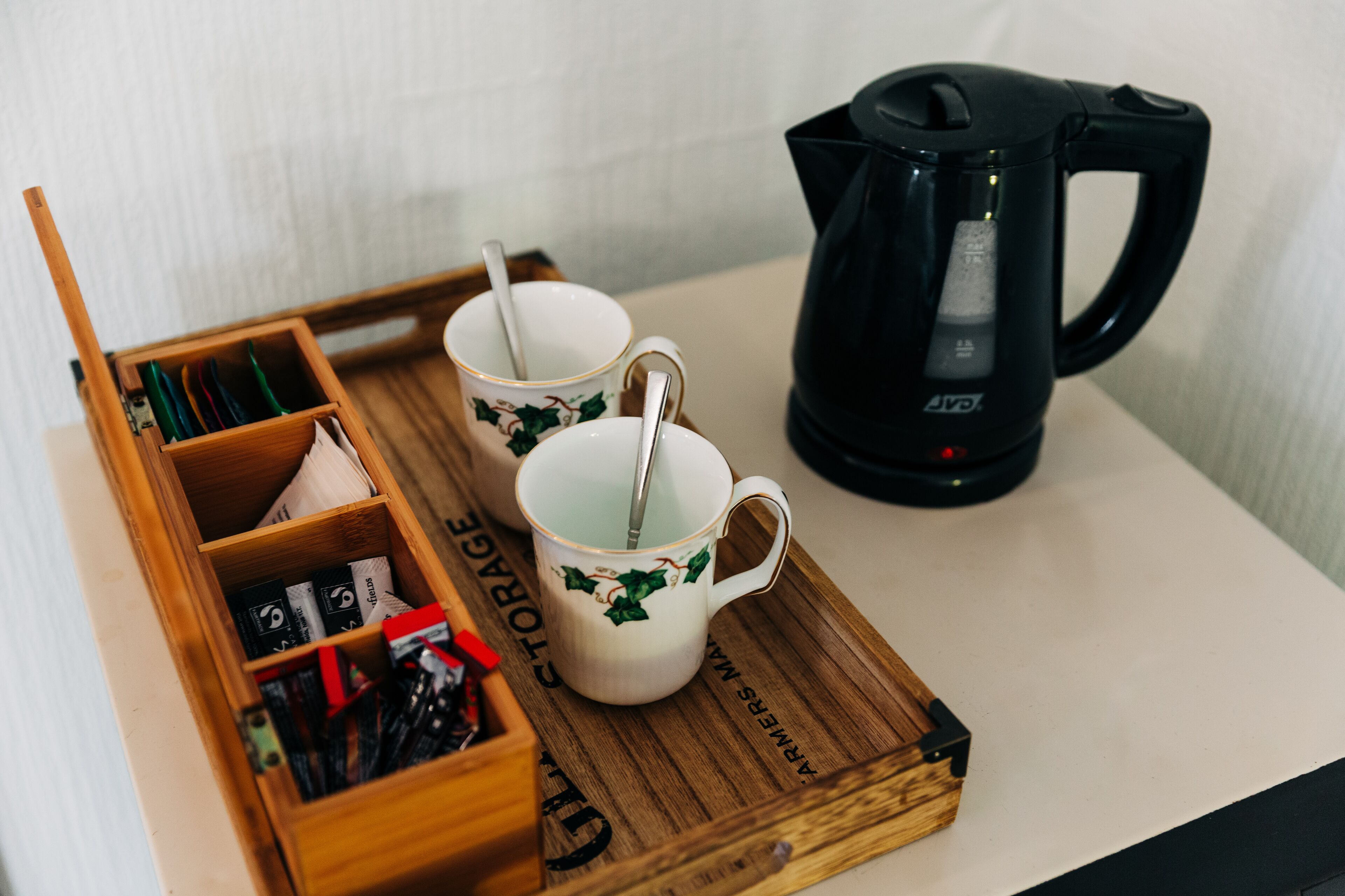A wooden tray with two white cups with green designs, a black electric kettle, and a storage box with various items.