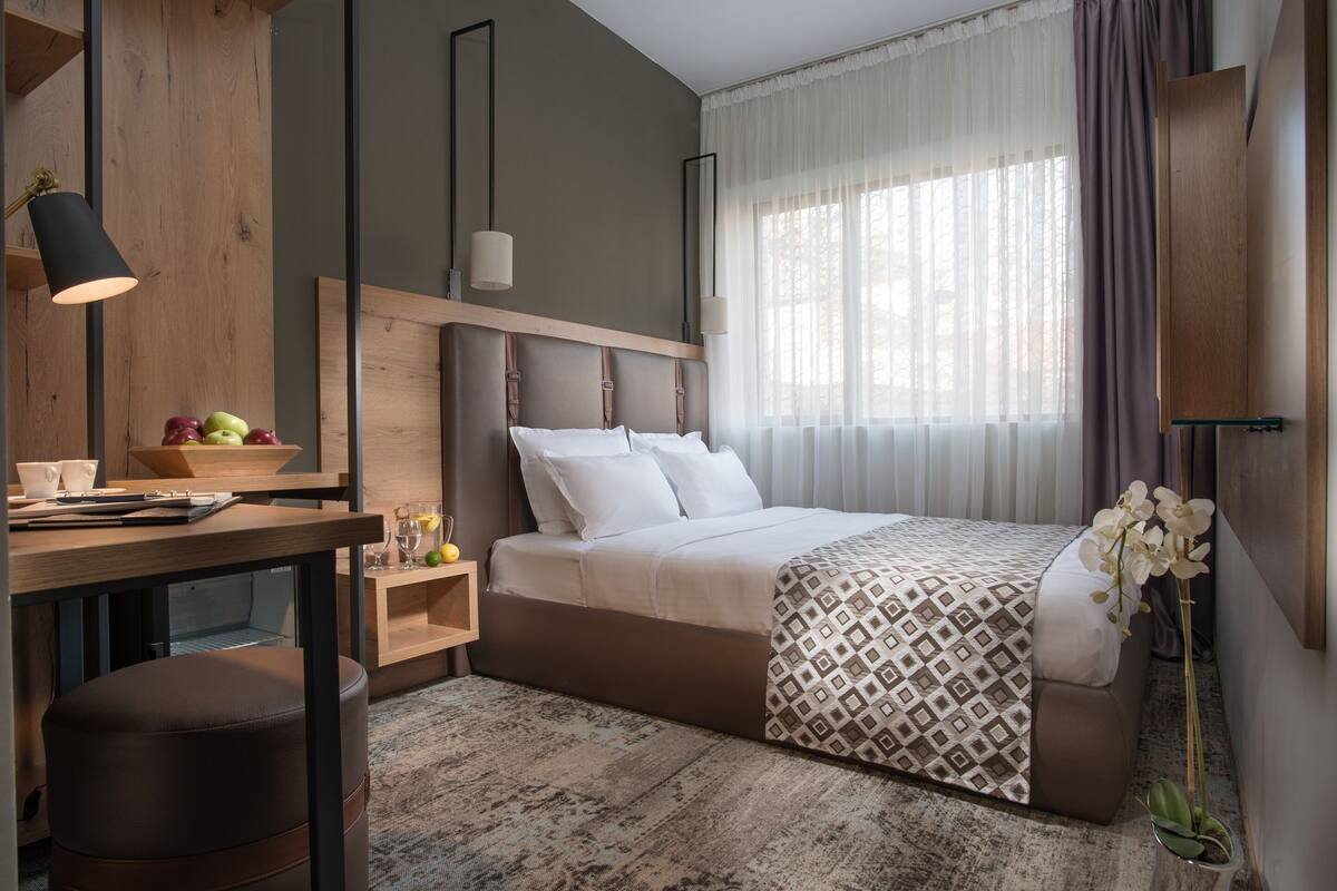A modern bedroom with a wooden headboard, a large bed with patterned bedding, a desk with a lamp, and a nightstand with a bowl of fruit.