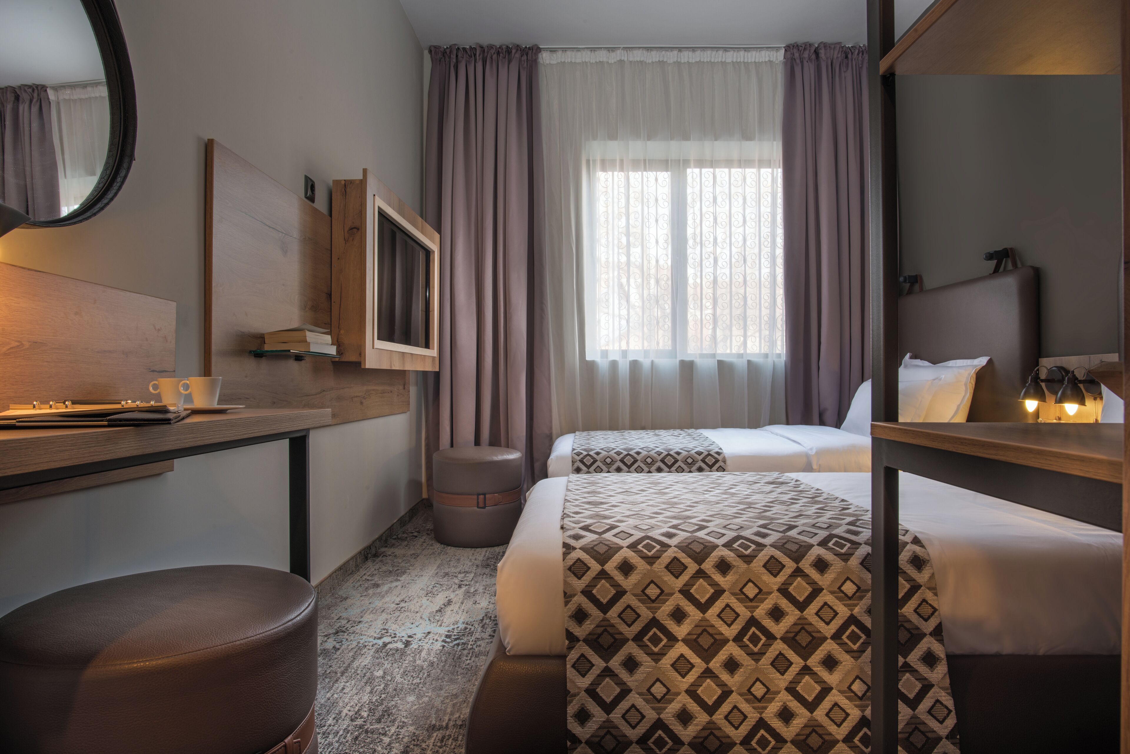 Een hotelkamer met een bed, een bureau met stoel, een spiegel, een raam met gordijnen en een klein krukje.