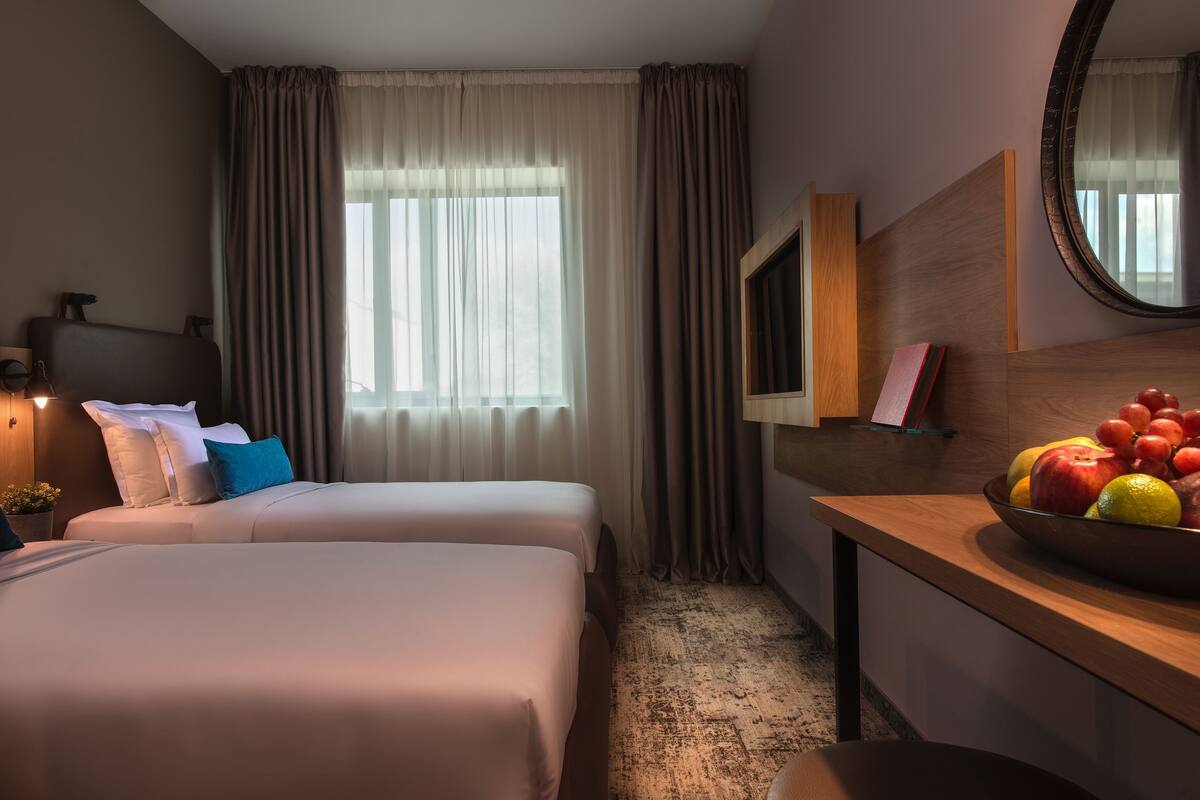 A hotel room with two beds, a desk with a fruit bowl, a mirror, and a window with curtains.