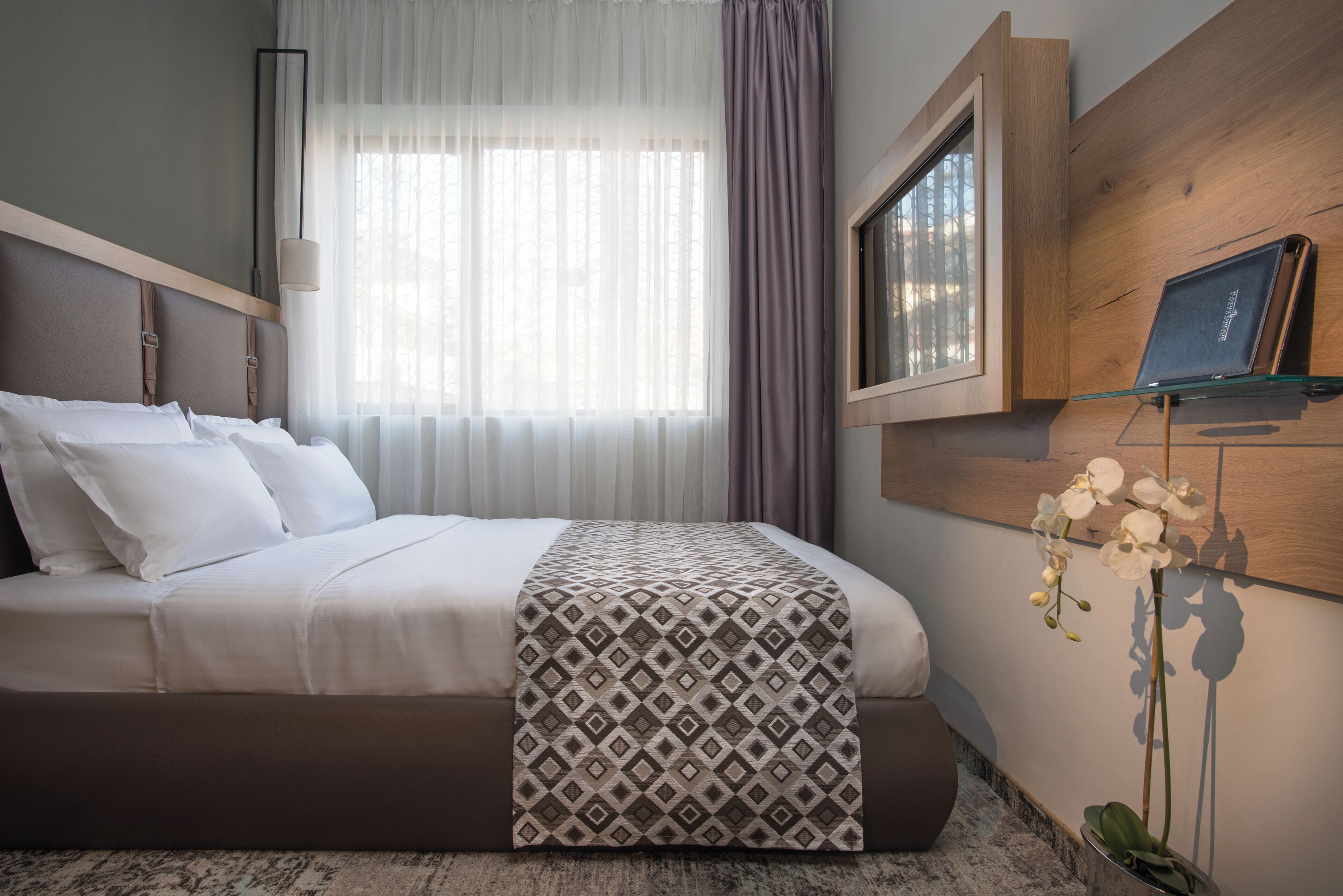 Habitación de hotel con cama, ventana con cortinas, televisor montado y orquídea en maceta.