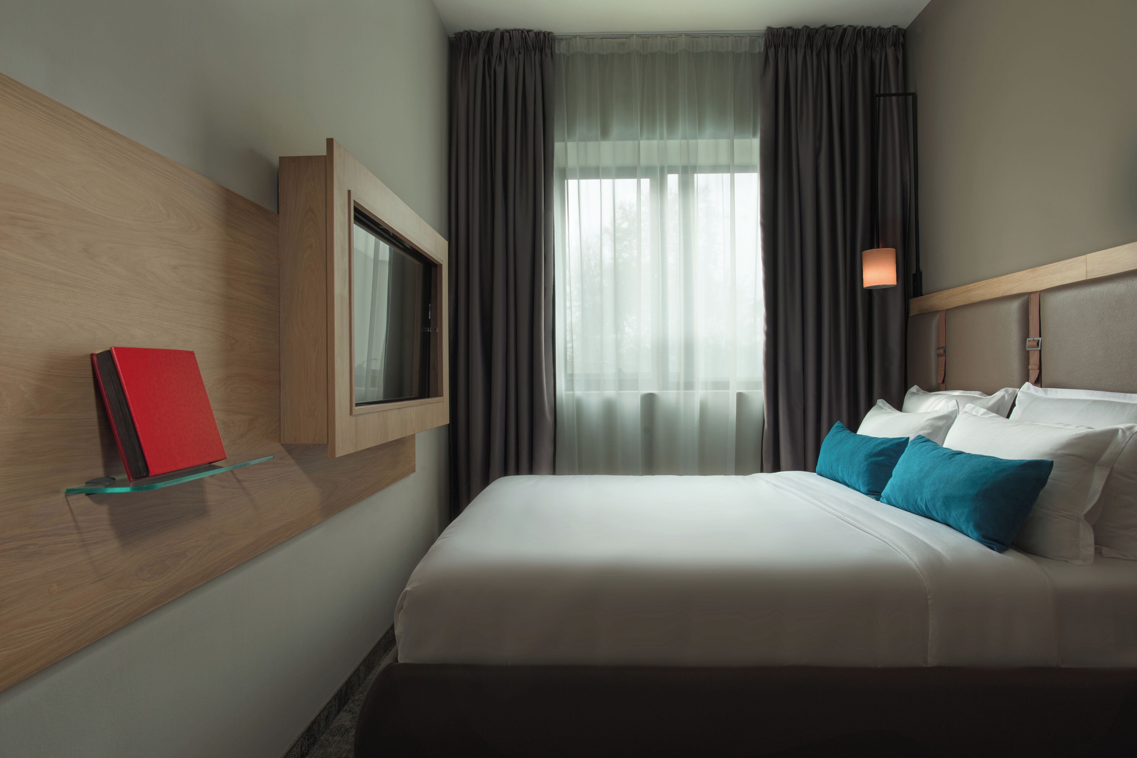 Habitación de hotel con cama, televisión montada en la pared, ventana con cortinas y mesita de noche con un libro rojo.