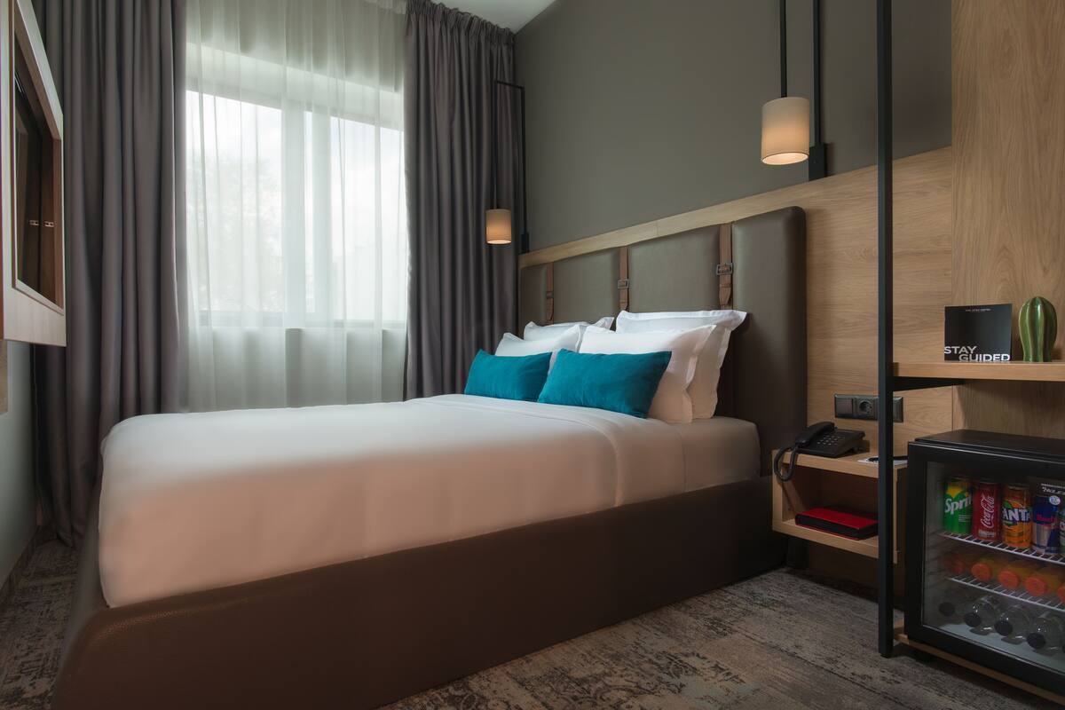 A modern hotel room with a large bed, a bedside table with a lamp, a minibar, and a view of the outdoors through sheer curtains.