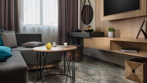 A modern living room with a grey sofa, a round wooden table with a yellow fruit bowl, a potted plant, and a mounted TV. - The Stay by Aston (Plovdiv)