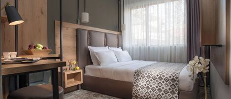 A modern hotel room with a bed, bedside table, lamp, and a view of the outdoors through sheer curtains.