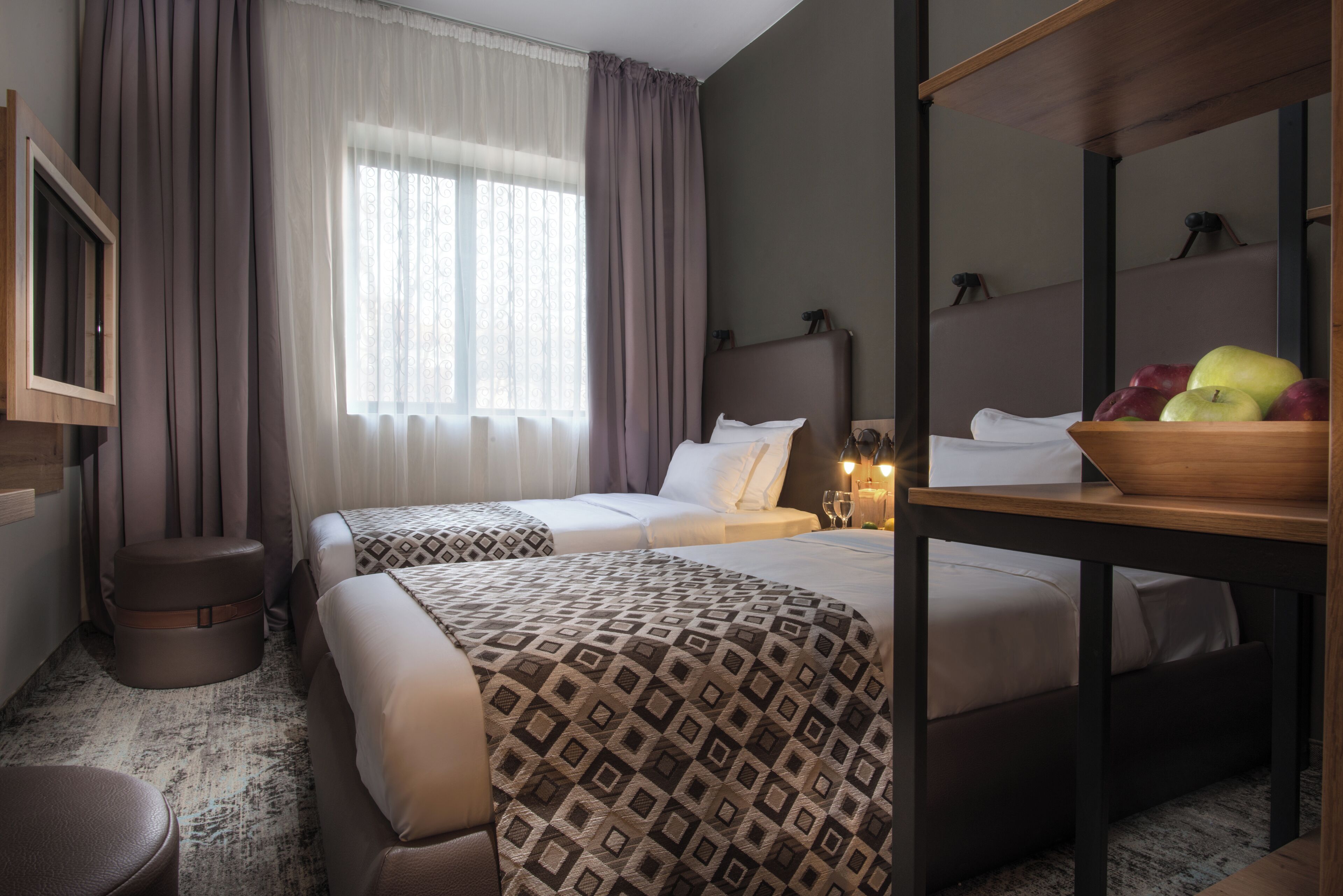 Et hotelværelse med to senge, et træsidebord med frugt, et spejl og et vindue med gardiner.