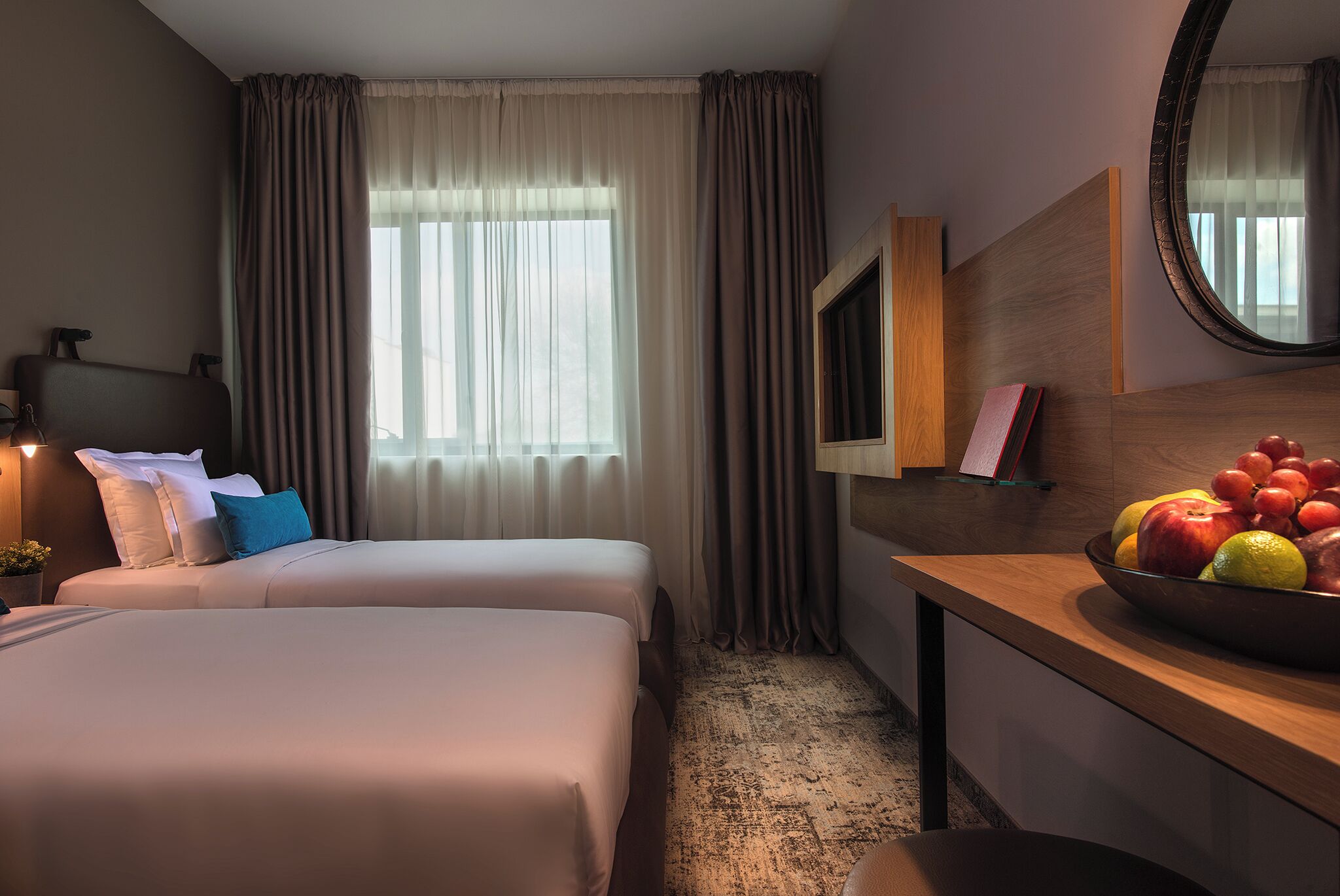 Une chambre d’hôtel avec deux lits, un bureau sur lequel se trouve un panier de fruits, une télévision et un miroir.