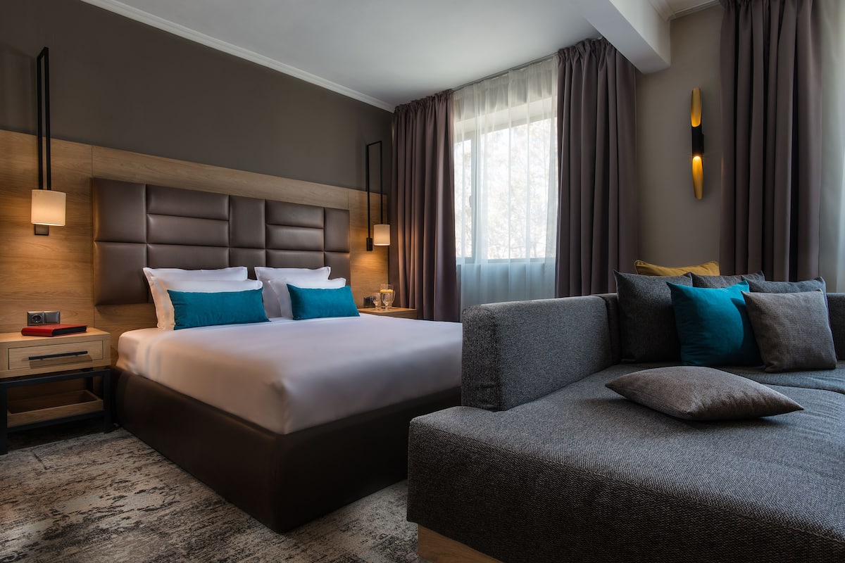 Ett modernt hotellrum med en stor säng, en soffa, ett nattduksbord och en väggmonterad lampa.