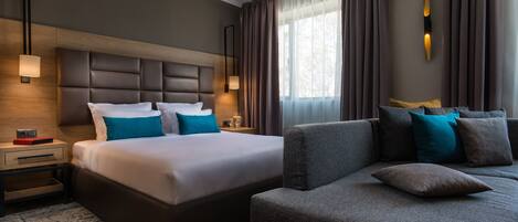 A modern hotel room with a large bed, a sofa, a nightstand, and a wall-mounted light fixture.
