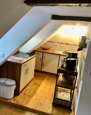 Une petite cuisine avec un plan de travail en bois, une cuisinière et un évier. On y trouve un meuble blanc à deux portes et une poubelle blanche.