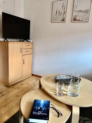 Een kamer met een houten kast, een televisie erbovenop, een ronde tafel met glazen en een boek, en ingekaderde foto’s aan de muur.