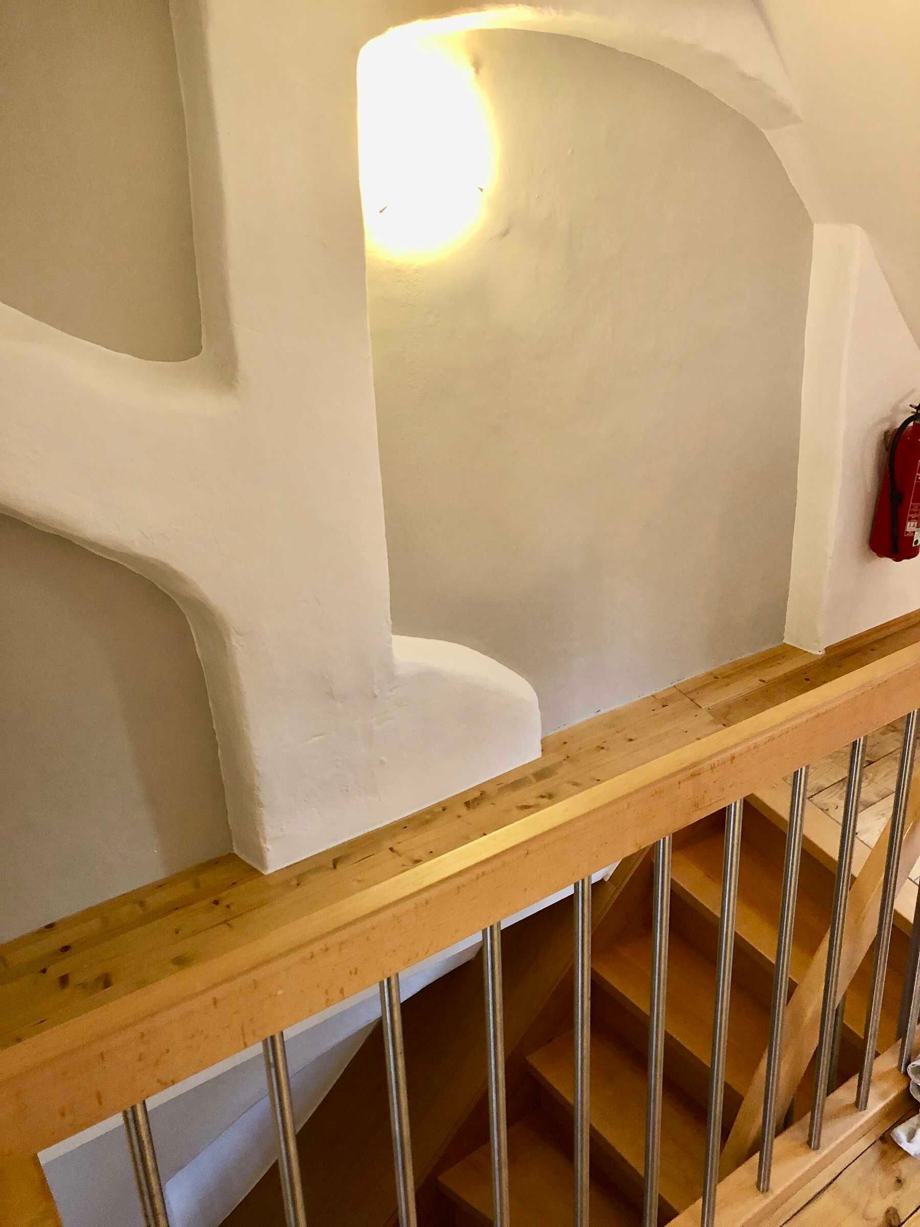 Un escalier avec une rampe en bois et des balustres en métal, menant à un palier avec un mur blanc et un extincteur rouge fixé sur ce mur.