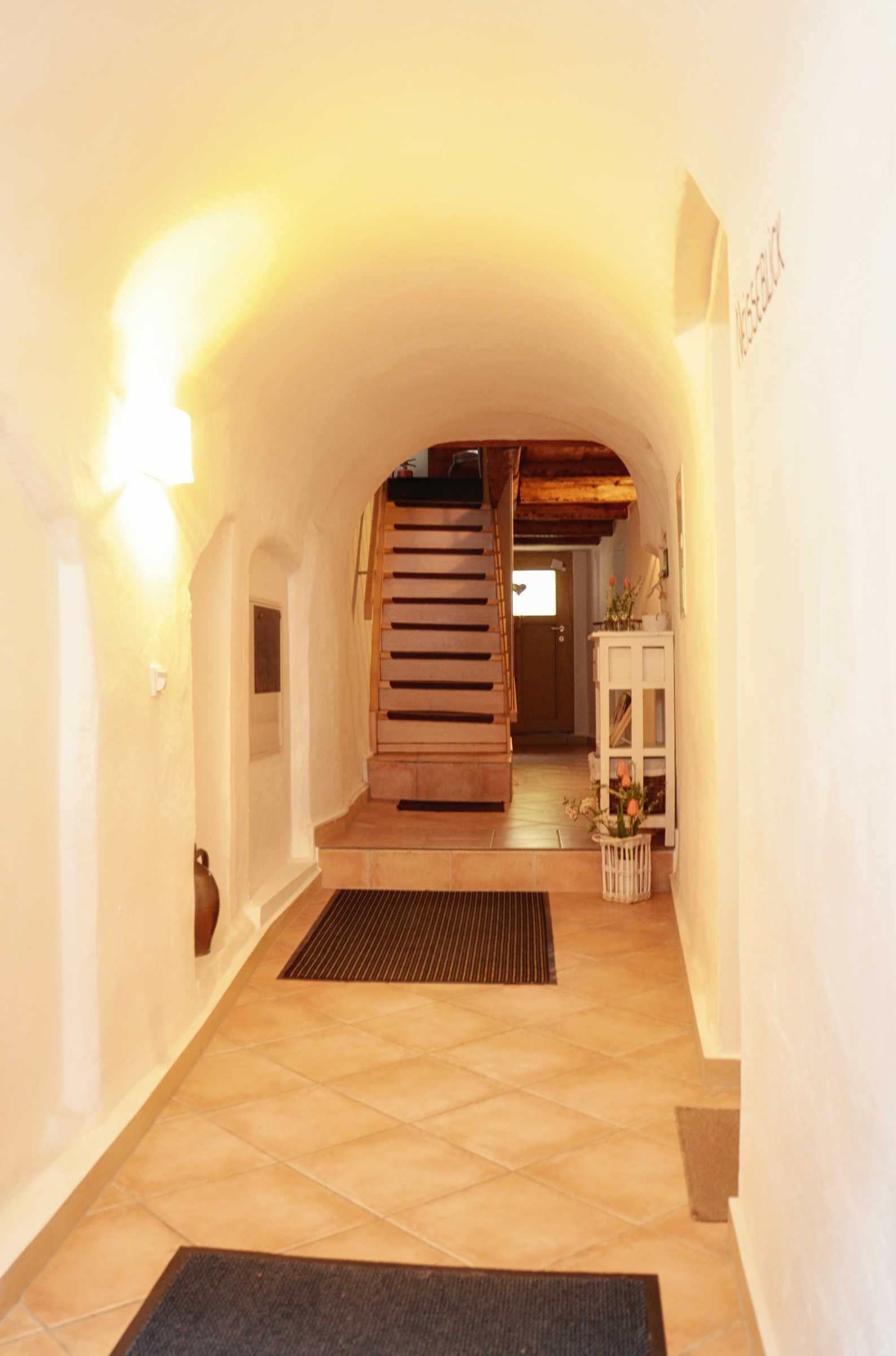 Un corridoio con una scala in legno, un armadietto bianco con fiori e una porta in fondo.