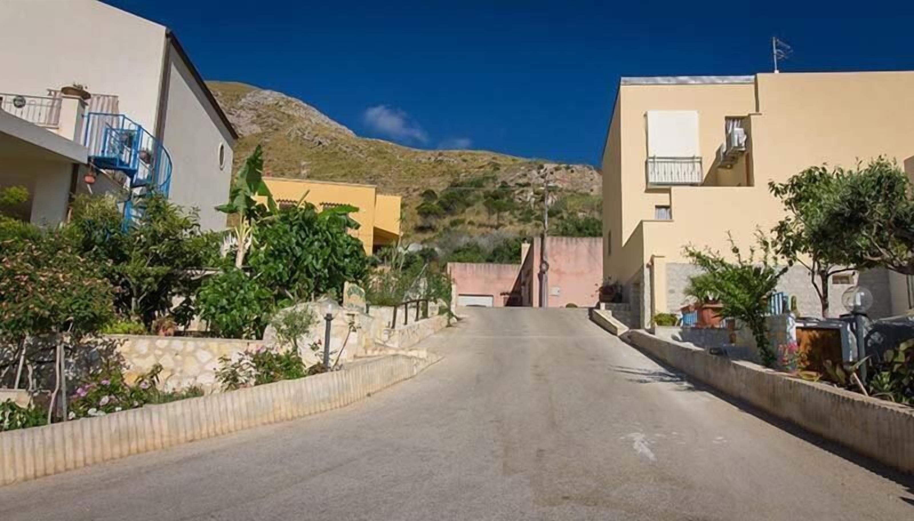 Una calle estrecha bordeada por edificios residenciales con balcones, un cielo despejado y un paisaje montañoso al fondo.