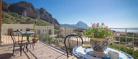 A terrace with a table and chairs, a potted plant, and a view of the sea and mountains.