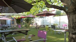Un patio al aire libre con mesas y sillas, rodeado de árboles y cubierto por sombrillas.