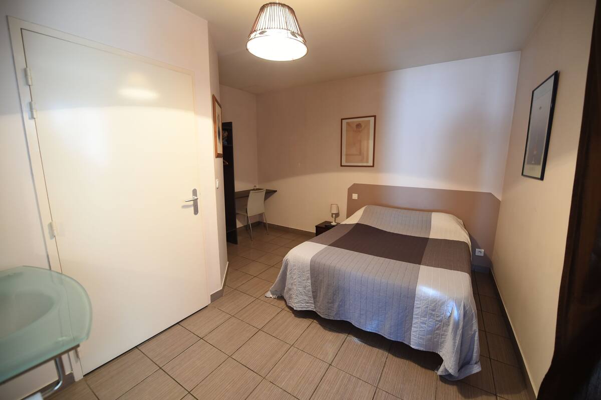 A bedroom with a bed, a chair, a bedside table, a lamp, and a framed picture on the wall.