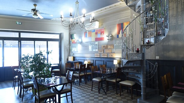 Comedor con candelabro, mesas y sillas de madera. Se ve una escalera con barandillas decorativas y una planta a la izquierda.