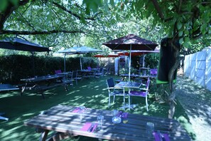 An outdoor patio area with tables and chairs, umbrellas, and a tree providing shade. - Le Juliano (Carbon-Blanc)