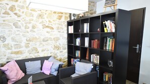 Una habitación con pared de piedra, una estantería negra llena de libros, un sofá negro con cojines y una pequeña mesa con un objeto rojo.